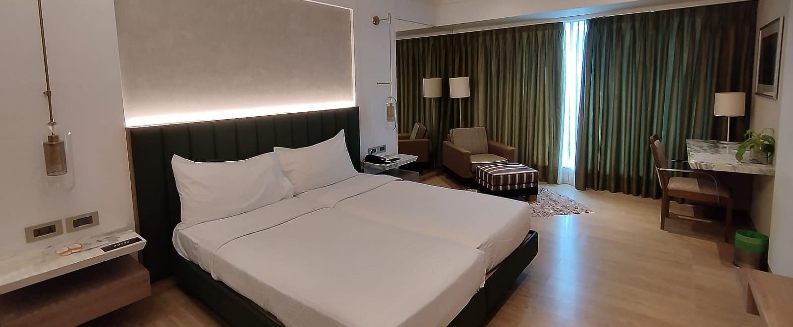 An executive bedroom in Mirage Hotel with well polished wooden floor, featuring twin beds, decorative head-rest, lounge chairs, and a large wall to wall window with closed drapes.