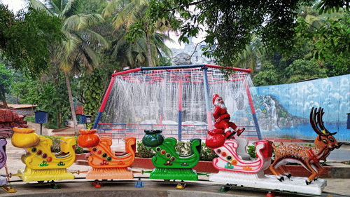 A colorful children's merry-go-round with animal-shaped seats and a Santa Claus figure, set against a backdrop of palm trees and a waterfall feature in an amusement park.