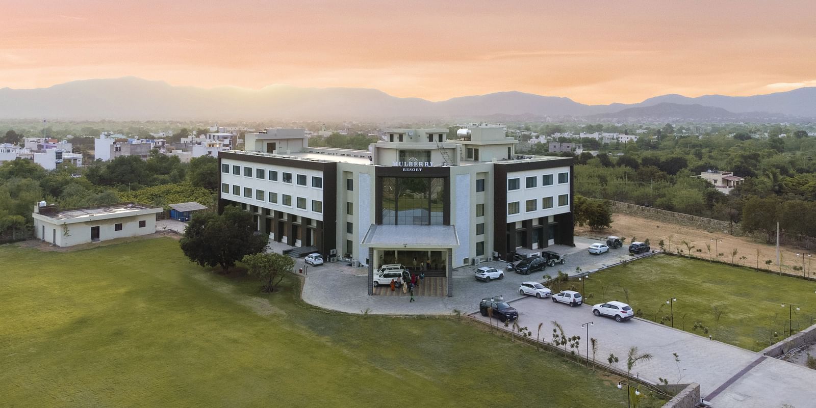 An aerial view of the Bamboo Saa Mulberry Resort, Pushkar at sunset or sunrise surrounded by green lawn and the backdrop of hills.