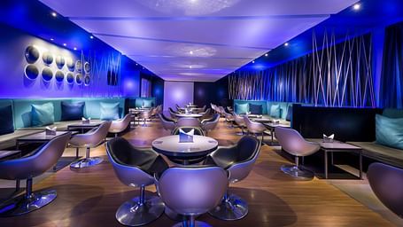 Modern restaurant interior with a blue and purple color scheme. The room features curved seating, round tables, and ambient lighting.