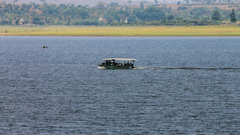 A river scene with a motorised boat carrying passengers across calm blue waters. The far shore has a grassy riverbank with scattered trees and hills in the background under a hazy sky.