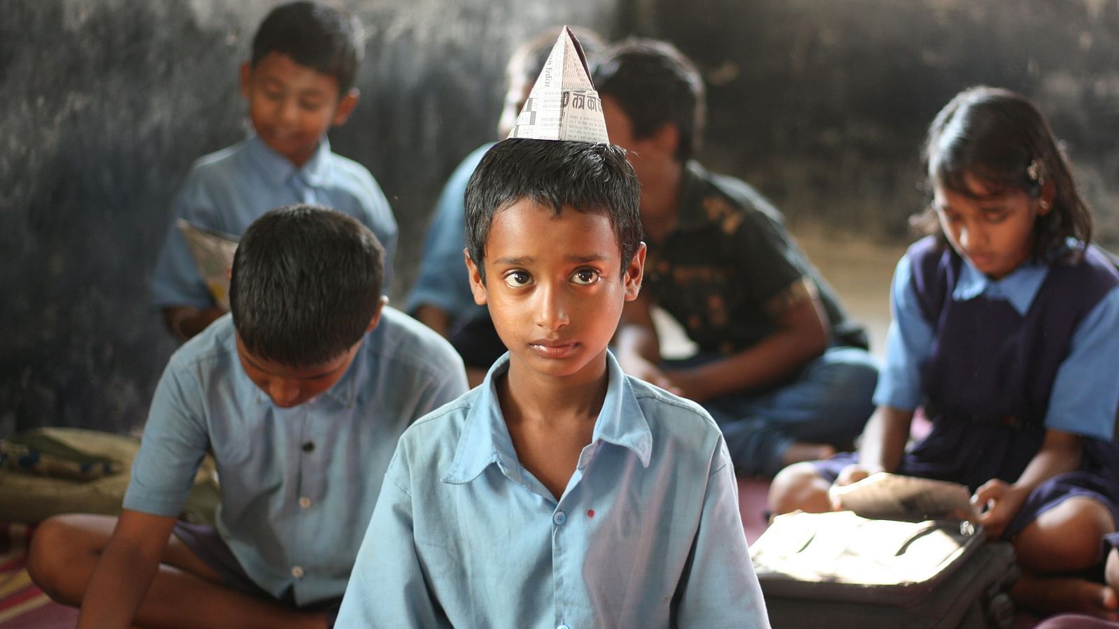 Front view of a young boy in a class wearing a paper hat with children in the background.