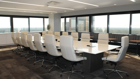 A modern conference room featuring a long rectangular table surrounded by white office chairs next to large windows.