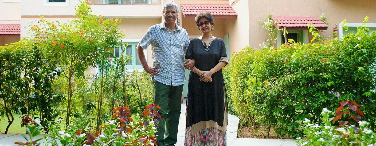 Two people with inviting smiles standing on a pathway bordered by plants with a background of the Karma Chalets  building.