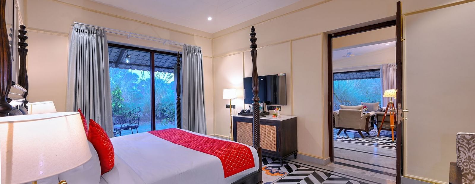 Premium Suite Cottage with a dark wood four-poster bed featuring red accents, black-and-white chevron flooring, a seating area, a wall-mounted TV, and an open doorway leading to another room - Singhvilas, Ranthambore