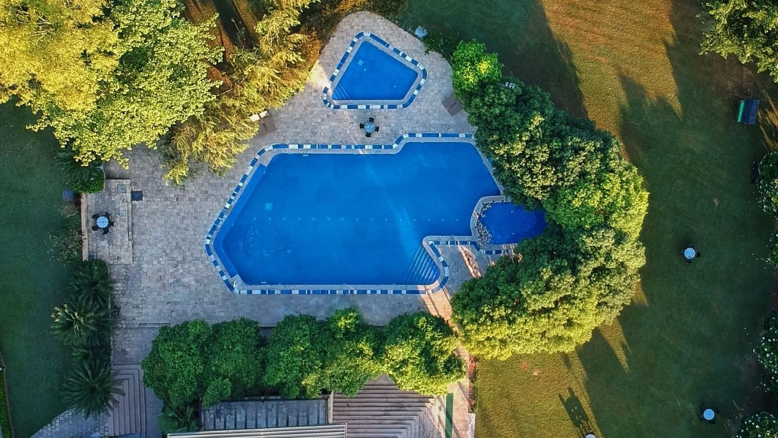 A drone shot of a huge swimming pool at Ivory Destinations surrounded by lush green trees and lawn.