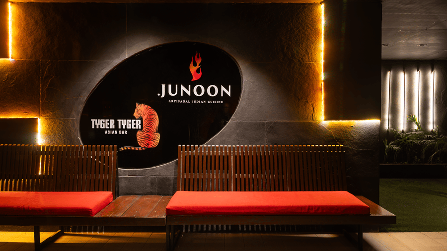 A restaurant entrance with a signboard reading 'JUNOON' above a bench and yellow lights on the wall at Infiniti Hotel, Indore.