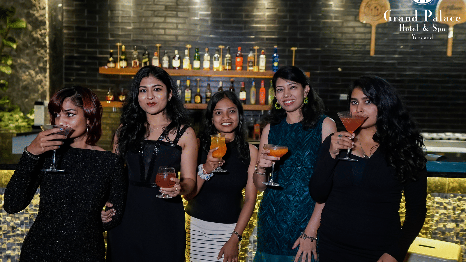 Group of guests holding drinks at the bar in Grand Palace, Yercaud with bottles arranged on the wall and garden lighting in the background.