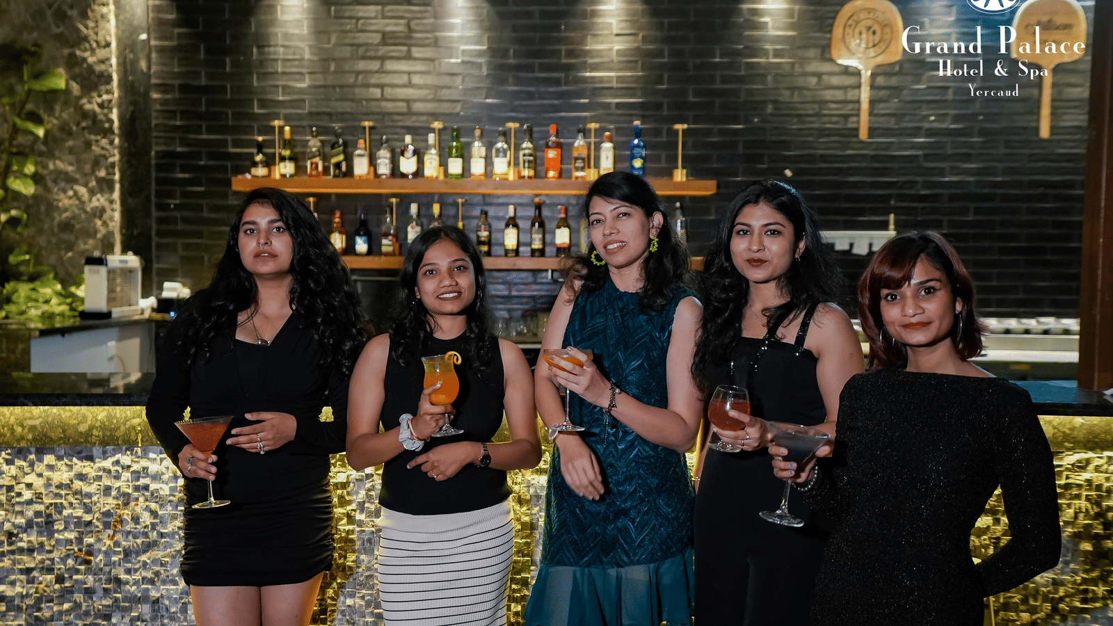 Guests standing at the bar counter in Grand Palace, Yercaud holding glasses with illuminated bottle display and green wall behind.