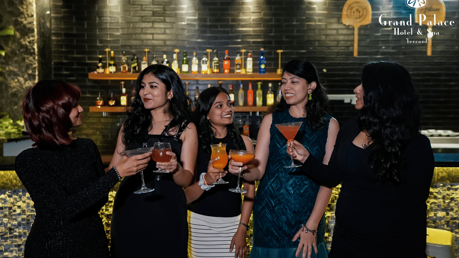 Guests gathered near the bar counter at Grand Palace, Yercaud holding drinks with wall-mounted bottle shelves in the background.