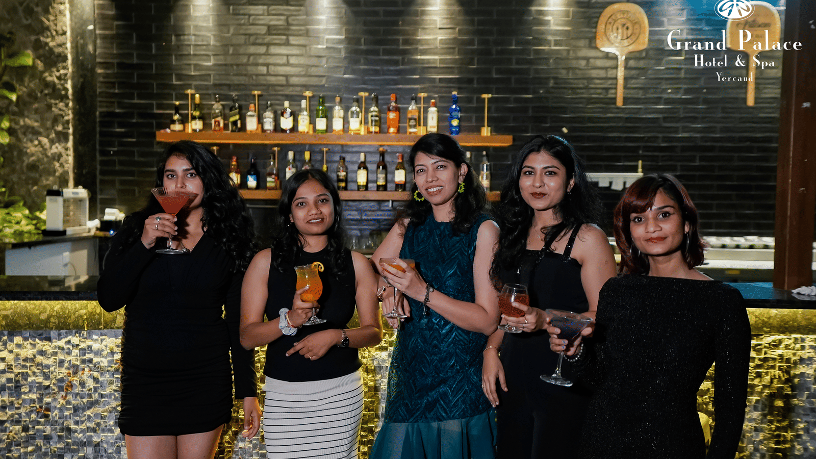 Group of ladies at the bar in Grand Palace, Yercaud holding glasses near the counter with illuminated bottle rack and wall lights.