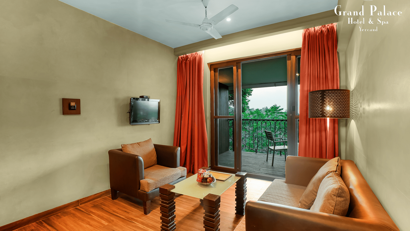 Living room at Grand Palace, Yercaud, with sofa, coffee table, chairs, large window, TV, ceiling fan and balcony view, furnished for guest comfort