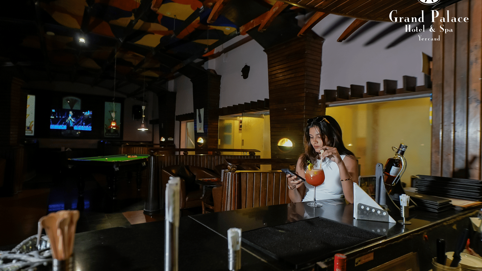 A lady at Cloud-9 Bar, Grand Palace, Yercaud preparing drinks near the bar counter with seating booths and billiards table.