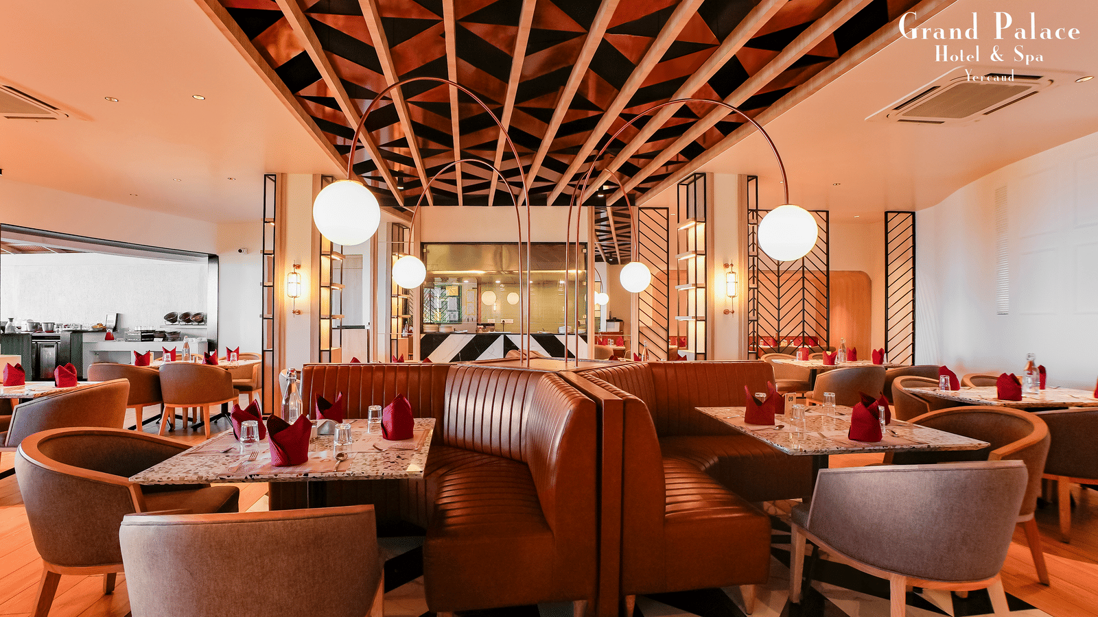 Indoor restaurant at Grand Palace, Yercaud showing dining tables, cushioned seating, ceiling beams, and wall lamps.