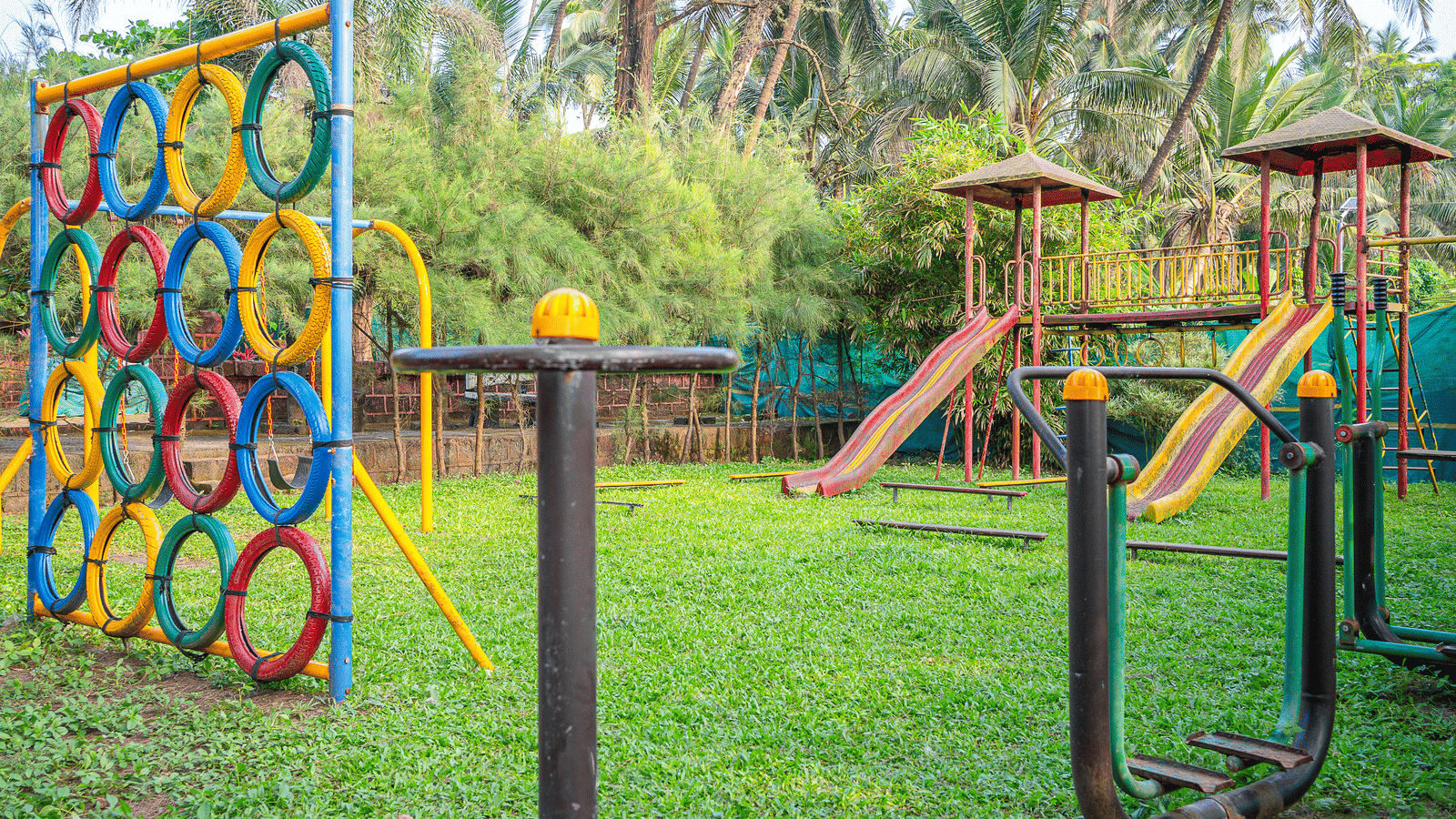 Playground with climbing structure and wooden play equipment, green grass, palm trees at Tranquil Beach Resort, Harihareshwar.