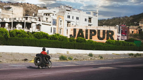Roadside view of a large Jaipur city sign with buildings, greenery, and a motorcyclist riding past under a cloudy sky.