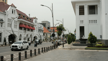 A street with with colonial-era buildings, a white car, and people walking on the sidewalk.