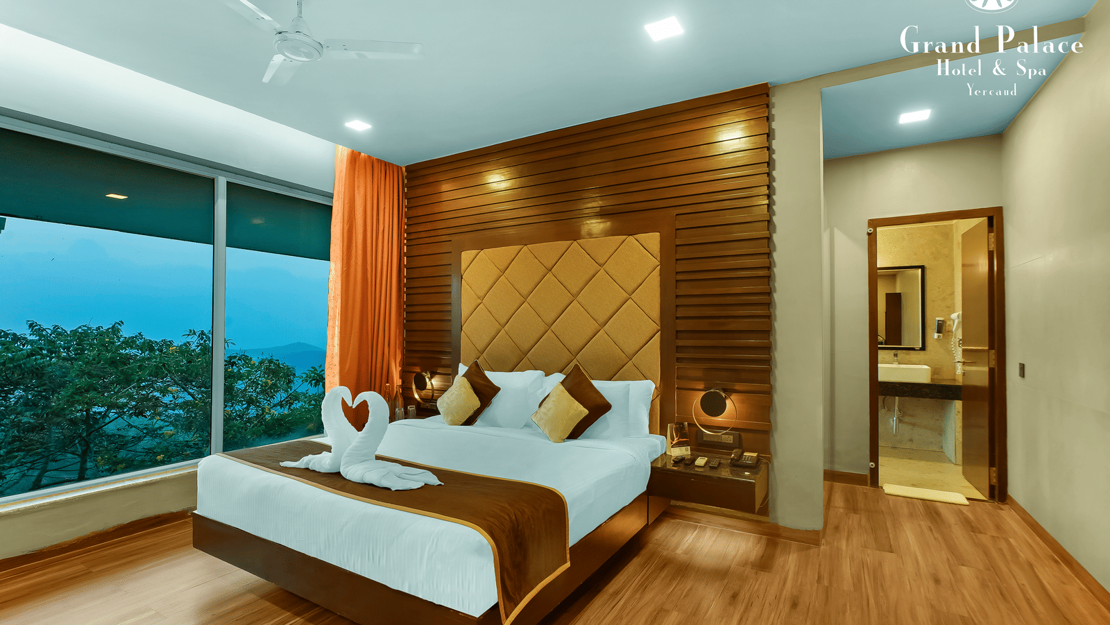 A corner angle Artist Suite at Grand Palace, Yercaud with twin beds, pillows, wooden headboard, curtains, floor lamp, large window, bedside table, and outside greenery visible