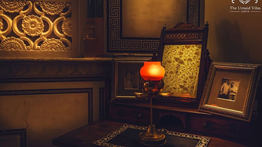 A dimly lit room at Umaid Vilas Jaipur featuring a decorative wall cutout, a lamp with an orange shade, and framed artwork.