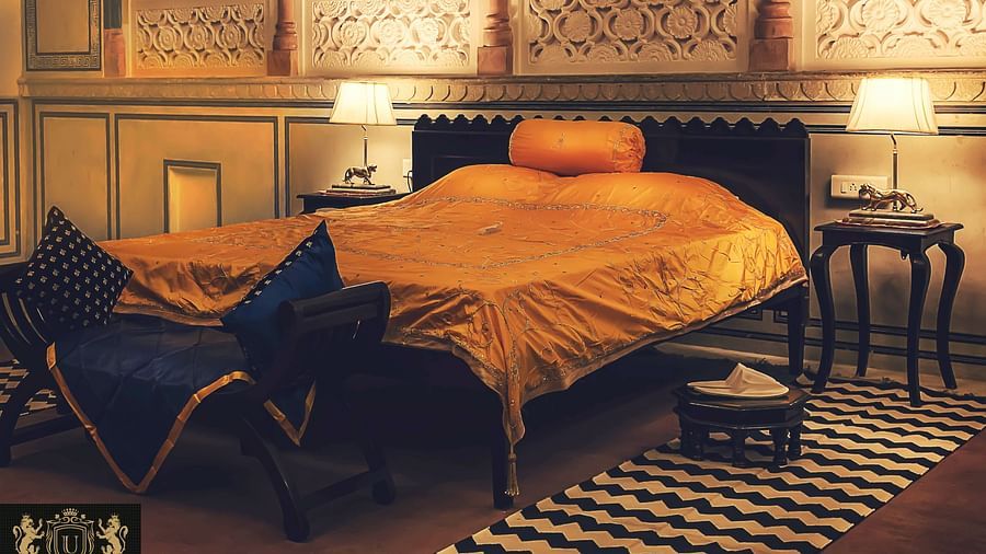 A bedroom at Umaid Vilas Jaipur with a bed dressed in golden fabric, two lamps, and decorative wall cutouts.