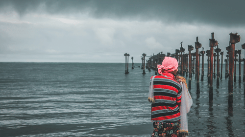 A man standing on seashore facing an old ruined structure of a port.