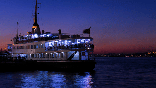 A view of a ferry during twilight hour with the lights on and people inside in view.