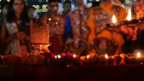 Close-up of lit diyas and offerings at a ceremonial prayer, with worshippers blurred in the background.