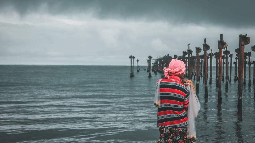 A man standing on seashore facing an old ruined structure of a port.