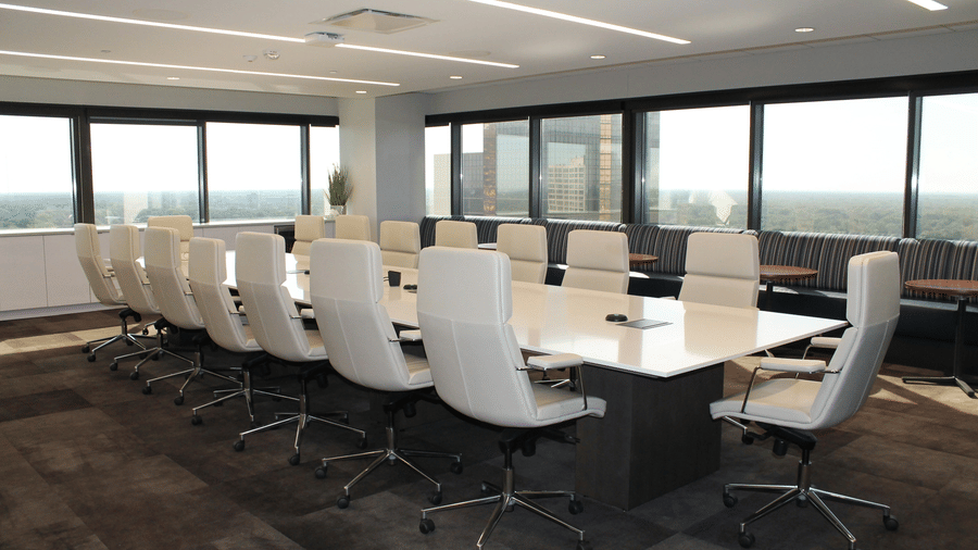 A brightly lit corporate boardroom featuring a long white table, high-back white office chairs and large windows lining the back wall