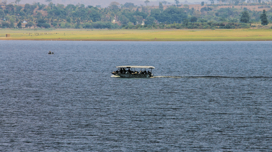 A river scene with a motorised boat carrying passengers across calm blue waters. The far shore has a grassy riverbank with scattered trees and hills in the background under a hazy sky.