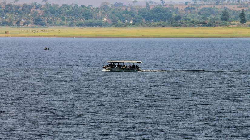 A river scene with a motorised boat carrying passengers across calm blue waters. The far shore has a grassy riverbank with scattered trees and hills in the background under a hazy sky.