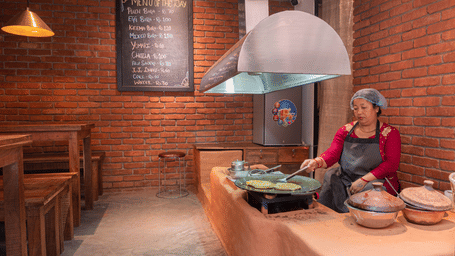 Woman cooking in a rustic brick kitchen at The Nanee, Bhaktapur, Nepal. stirring a large pot as warm light highlights traditional cookware.