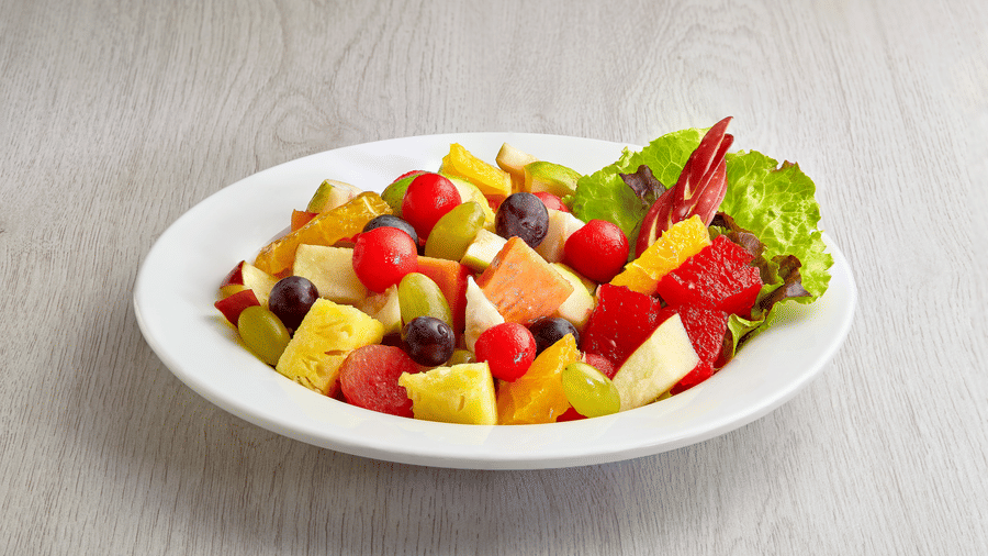 White round plate with a seasonal fruit salad of cherry tomatoes, grapes, watermelon, orange, pineapple, papaya and lettuce on a wooden surface.