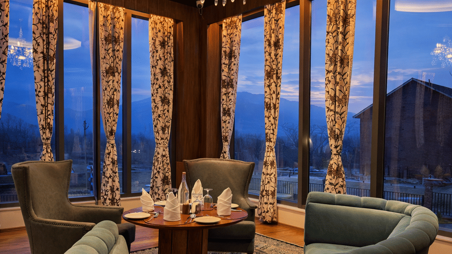 A dining space with circular seating arrangement at Mountnoor Sarovar Premiere. The space includes a curved sofa, armchairs, and a chandelier, all set against tall windows overlooking the valley at dusk.