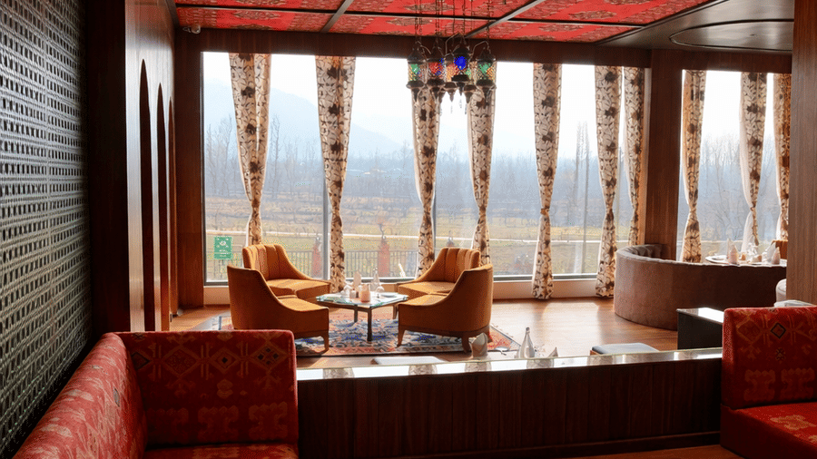 A dining space at Mountnoor Sarovar Premiere featuring a long built-in bench with patterned upholstery. The room opens up to a brighter area with armchairs and windows framed by patterned drapes.