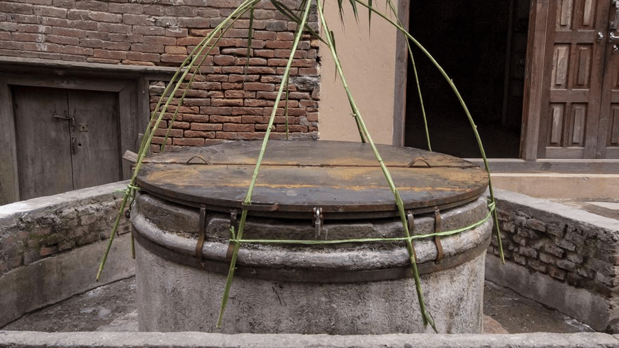 An ancient circular brick water well decorated with a ceremonial green leaf wreath outside a heritage building in Bhaktapur, Nepal.