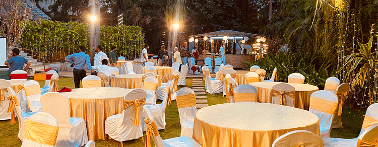 Outdoor event setup with round tables covered in white and gold cloths, surrounded by chairs under string lights.