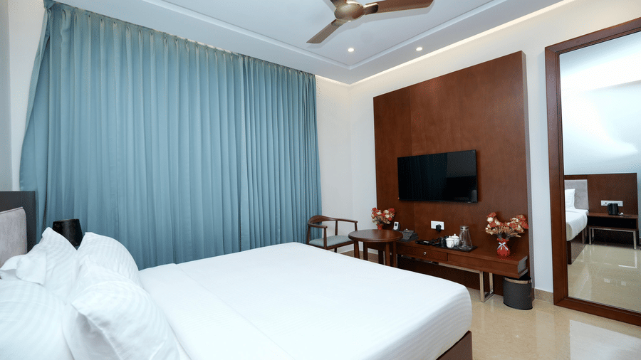 The Deluxe King Room of Le Royale Palace with a neatly made double bed, a flat-screen TV mounted to the wall, a large window with draped curtains, and wooden interiors