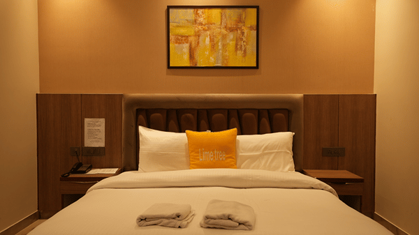 A centred shot of a neatly dressed double bed with a wooden headboard and a colourful abstract painting above.