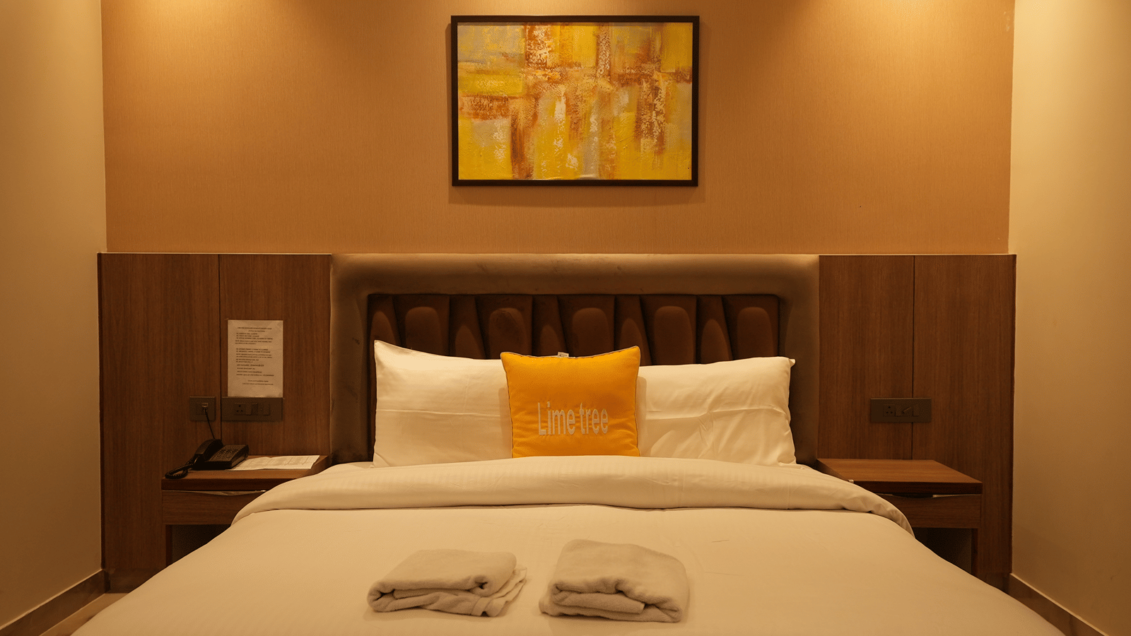 A centred shot of a neatly dressed double bed with a wooden headboard and a colourful abstract painting above.