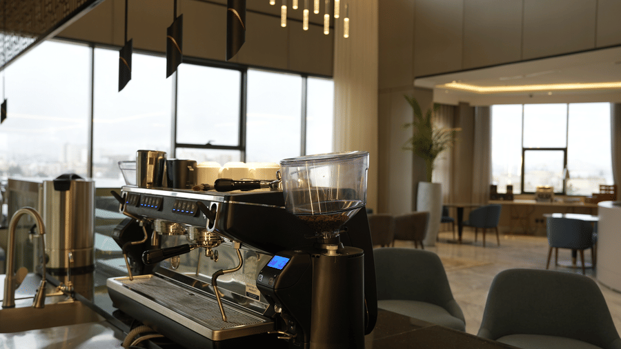 Counter of Grand Iva Cafe at Grand Iva Hotel, Abha, featuring a coffee machine, hanging ambient lighting, and large glass window showcasing the city's views.