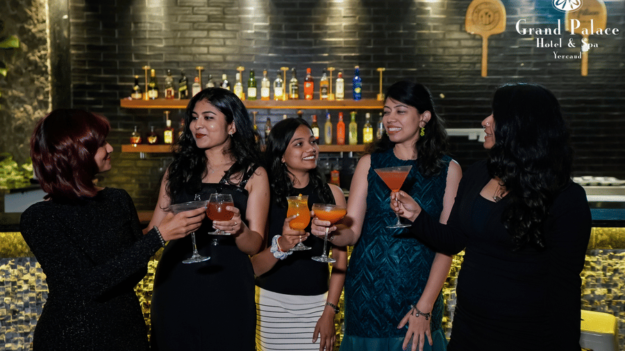 Guests gathered near the bar counter at Grand Palace, Yercaud holding drinks with wall-mounted bottle shelves in the background.