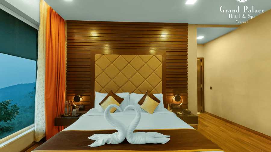 King bed at Grand Palace, Yercaud with towel art, pillows, wooden accent wall, bedside lamps, orange curtains, large window, and visible partition door.