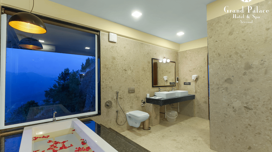 Bathroom at Grand Palace, Yercaud with bathtub, rose petals, separate shower section, basin, mirror, glass divider, and exterior night landscape visible through window .