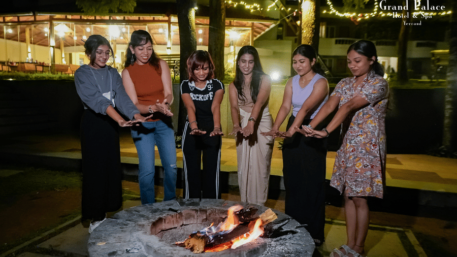 People standing by campfire at night at Grand Palace, Yercaud, with logs, firepit, outdoor setting, vegetation, lit faces and group reflection on experience.