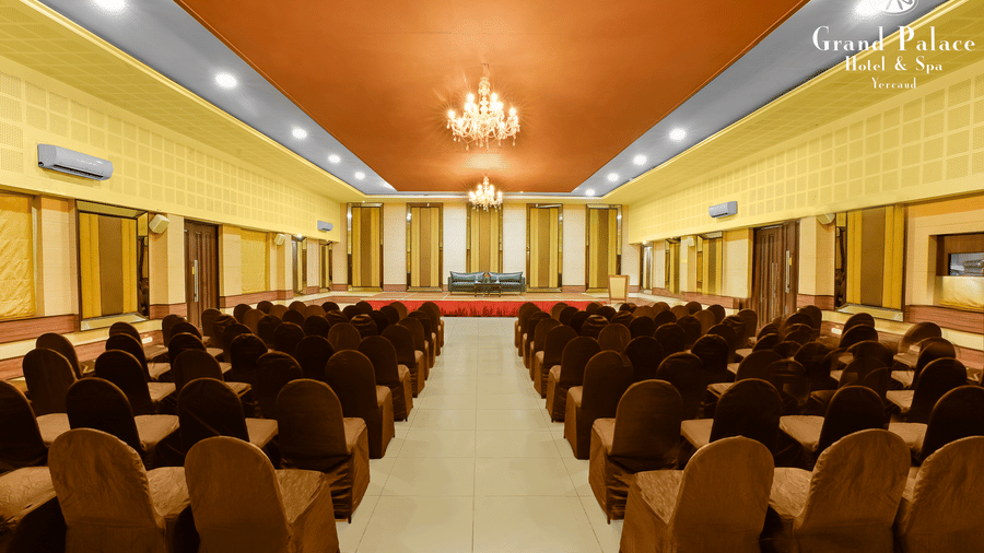 Grand I hall at Grand Palace, Yercaud with rows of chairs, centre aisle, chandeliers, projector screens and high ceiling suitable for meetings or seminars.