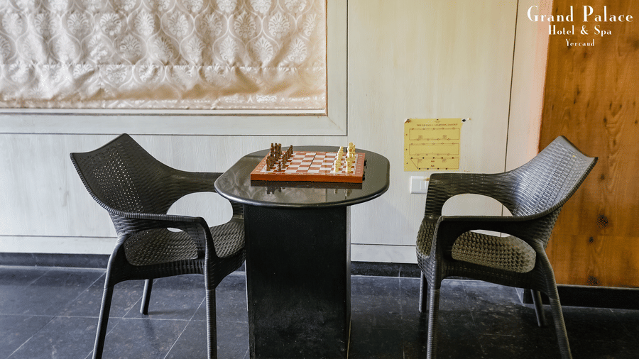 Table for two with a chessboard at Grand Palace, Yercaud, set near a curtained window with artwork and chairs, creating a cozy indoor dining space.