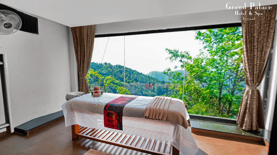 Spa at Grand Palace, Yercaud with bed, wooden floor, curtains, single bed, TV, window, green scenery visible outside the window.