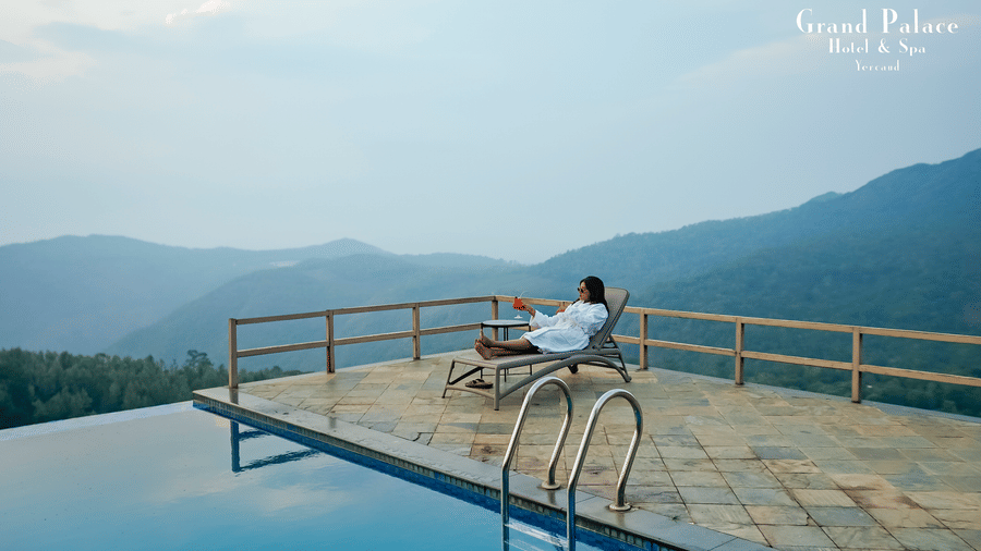 Woman sitting atop infinity pool deck at Grand Palace, Yercaud, overlooking surrounding forested hills, water, railing and sky view .