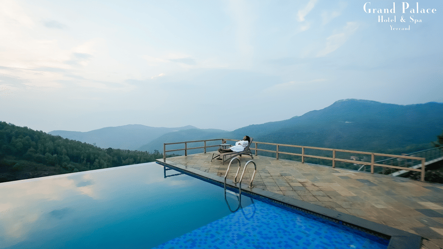 View of infinity pool at Grand Palace, Yercaud with deck, railing, distant hills, water surface, platform and wide sky above.
